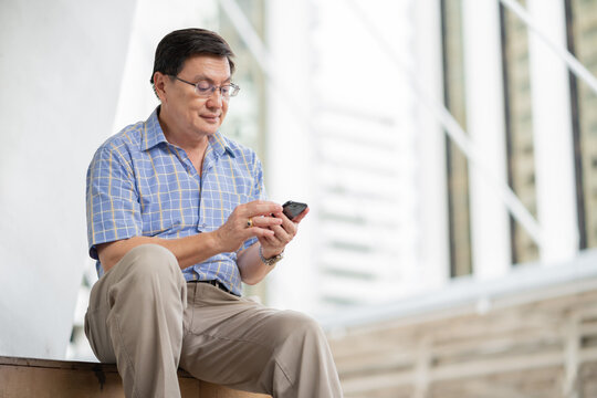 Asian senior man little smile looking picture on smartphone while sitting relax outside of the city