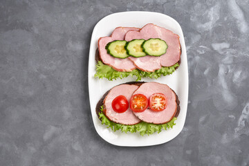 Two sandwiches with ham, cucumber and cherry tomato on a white plate on a gray background. Copy space.