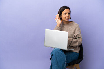 Young mixed race woman sitting on a chair with laptop isolated on purple background listening to something by putting hand on the ear