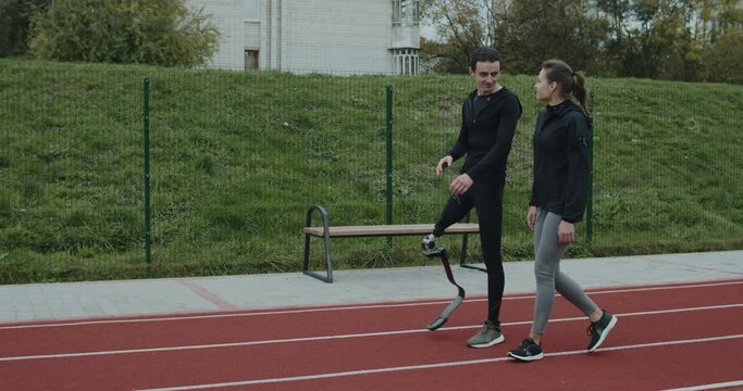 Side view of cheerful man with prosthetic running blade talking to woman outdoors. Male sportsman with amputed leg chatting with female persom and smiling while walking at sports field.