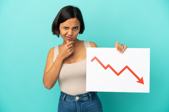 Young mixed race woman isolated on blue background holding a sign with a decreasing statistics arrow symbol and thinking