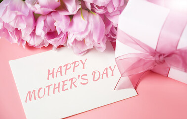 A bunch of pink tulips on a warm pink background with a card with HAPPY MOTHER'S text and a gift.
