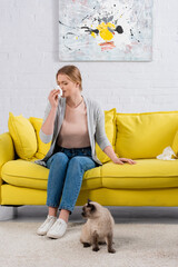 Woman with napkin sneezing near siamese cat on carpet