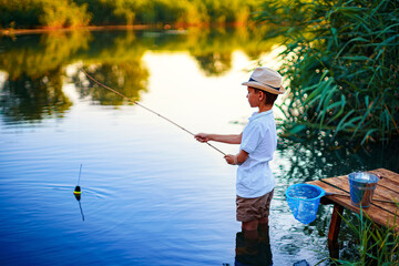 Little boy in straw hat fishing in lake at sunset.