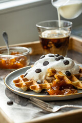 Homemade Belgian waffles with natural whipped cream, chocolate droplets and apricot jam,  glass of coffee , milk jug and jam on marble tray.