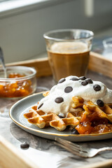 Homemade Belgian waffles with natural whipped cream, chocolate droplets and apricot jam,  glass of coffee , milk jug and jam on marble tray.