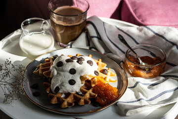 Homemade Belgian waffles with natural whipped cream, chocolate droplets and apricot jam,  glass of coffee , milk jug and jam on little round table near armchair .