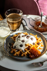 Homemade Belgian waffles with natural whipped cream, chocolate droplets and apricot jam,  glass of coffee , milk jug and jam on little round table near armchair .