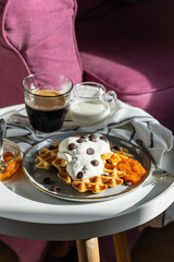 Homemade Belgian waffles with natural whipped cream, chocolate droplets and apricot jam,  glass of coffee , milk jug and jam on little round table near armchair .