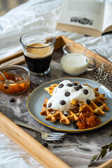 Breakfast in bed. Homemade Belgian waffles with natural whipped cream, chocolate droplets and apricot jam,  glass of coffee , milk jug and jam on marble tray.