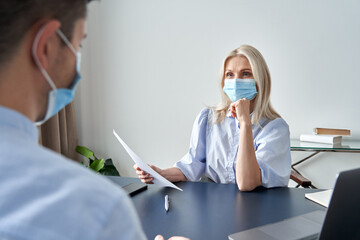 Female hr specialist manager, employer wearing face mask holding cv listening male candidate at job interview meeting. Hiring and employment during covid social distance safety concept.