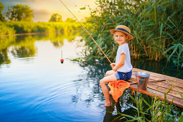 Little boy in straw hat sitting on the edge of a wooden dock and fishing in lake at sunset.