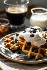 Homemade Belgian waffles with natural whipped cream, chocolate droplets and apricot jam,  glass of coffee , milk jug and jam on marble tray.