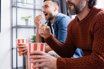 Interracial father and son watching tv while eating popcorn at home, blurred background