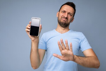 Dissatisfied handsome young unshaven brunet man wearing everyday blue t-shirt isolated over blue background holding and showing mobile phone with empty display for cutout looking at camera and saying