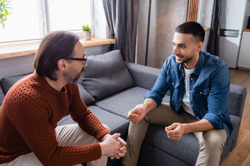 Young Hispanic man gesturing while talking to dad on sofa at home