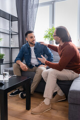 Fototapeta premium father touching shoulder of smiling hispanic son while sitting on sofa in living room