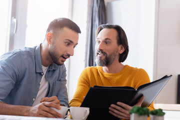 Amazed Hispanic man looking at photo album near father, blurred foreground