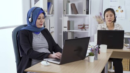 portrait Islamic expectant service agent sitting at desk with headphones is using the computer while smiling and talking to customer via phone call.