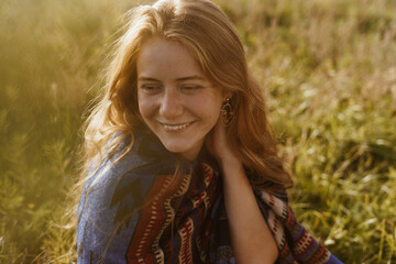 a sincere smile on a beautiful face with freckles at the red-haired girl in this cape sitting on the yellow steppe grass. High quality photo