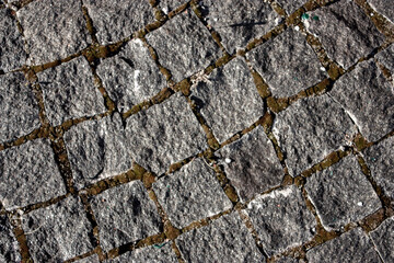 The sidewalk is paved with stones, top view. Grass and moss grow between the cobblestones on the pavement. Sharp edges of stones on a new pavement on a summer day.