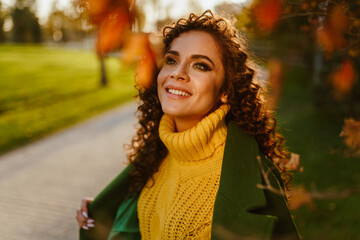 Smiling beautiful white-toothed smile curly girl with long dark hair enthusiastically looking up against the background of autumn leaves. High quality photo