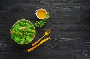 Fresh green vegetable salad with lettuce and carrot in bowl with orange knife and fork on dark wooden background, healthy vegan eating and dieting concept, top view with copy space