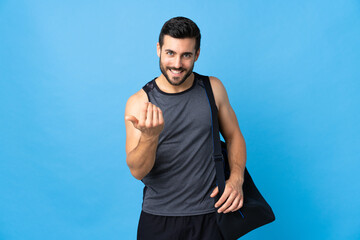 Fototapeta premium Young sport man with sport bag isolated on blue background inviting to come with hand. Happy that you came