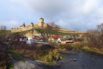 Fototapeta na wymiar Beautiful landscape photo of ancient Kamianets-Podilskyi fortress on the cliff against blue sky. Few residential houses under the hill. Smotrych River in the foreground. Travel and tourism concept