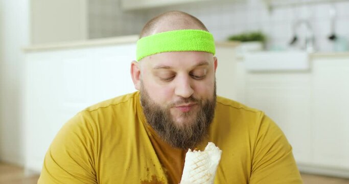 Portrait Of Sweaty Bearded Fat Man In Yellow Wet Sportswear Is Eating Danar In Kitchen At Home. Enjoying Junk Food. Trying Loosing Weight. Humor, Mem, Parody, Fun Behaviour. Addiction Of Food.