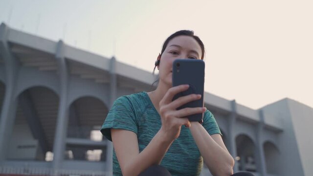 asian athlete woman wearing headphone using mobile smart phone sitting on stair at sport club stadium in evening sunset. female in sportswear  using online application tracking activity and music.