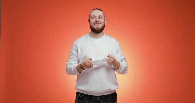 Young friendly bearded man with tail in hairs in sweater and jeans takes off medical mask and makes a beed breath on red background, front view. Covid-19, coronavirus pandemic, epidemic, quarantine.