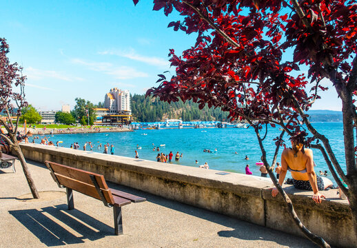 A Woman Relaxes On A Barrier Wall At The City Beach And Park Along The Shores Of Lake Coeur D'alene, Idaho On A Busy Summer Day.