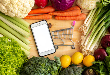For easy grocery shopping, order online and buy vegetables and eat healthy. Isolated mobile phone and supermarket cart with vegetables.