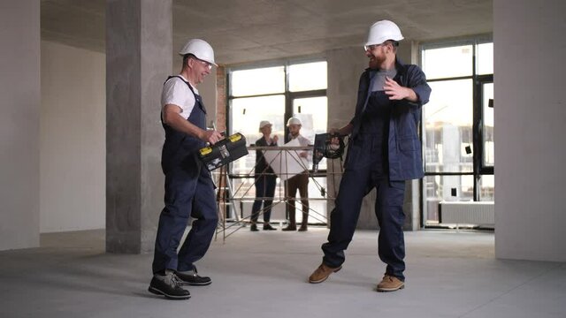 Happy builders in protective helmets and glasses, with screwdriver and repair toolbox in hands enjoying funny dance in apartment under renovation while architects planning interior space on background