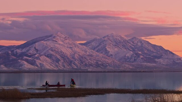 People in canoes during sunset on Utah lake against snow capped mountains as one splashes water with paddle trying to get the others wet.