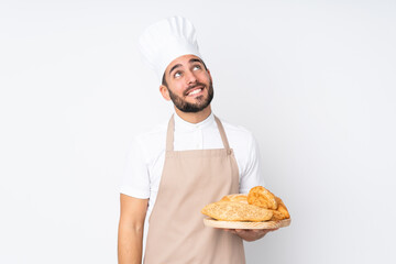 Male baker holding a table with several breads isolated on white background laughing and looking up