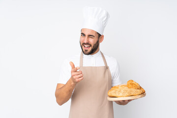 Male baker holding a table with several breads isolated on white background points finger at you