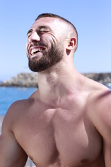 Hombre musculado sin camiseta riendo mientras disfruta de un día de sol en la playa. Concepto de emoción, verano y diversión