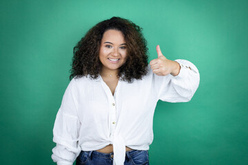 Young african american girl wearing white shirt over green background success sign doing positive gesture with hand, thumb up smiling and happy. cheerful expression