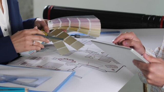 Close-up of designers hands over paper blueprint of house layout, woman choosing colors for painting using color palette, man working on tablet. Two designers planning color scheme of interior