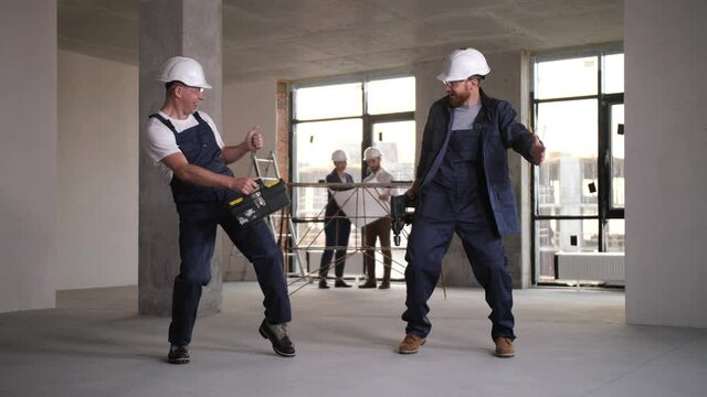 Joyful male workers in helmets, protective glasses and repair tools having fun during dance among empty space of renovated apartment. Funny builders dacing while designers working on background