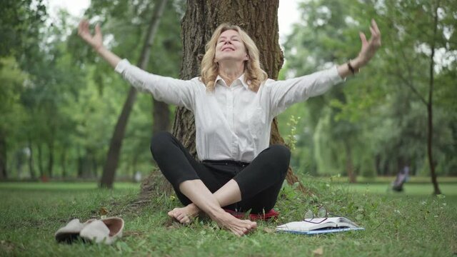 Portrait of confident middle aged barefoot businesswoman taking off eyeglasses and sitting in lotus pose in summer park. Wide shot of beautiful elegant woman meditating outdoors. Life balance.