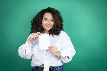 Young african american girl wearing white shirt over green background smiling and showing blank notebook in her hand