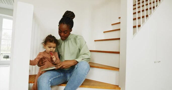 Smiling young African mother reading a story to her cute little daughter while sitting on some stairs at home