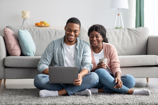 Happy black lovers sitting on carpet at home, using laptop