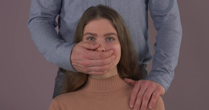 Zoom-in portrait of abused Caucasian woman. Unrecognizable Caucasian man covering her mouth with hand. Domestic violence. Abuse in relationship.