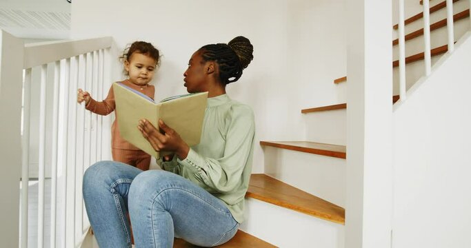 Smiling young African mother reading a story to her adorable little girl while sitting on stairs at home