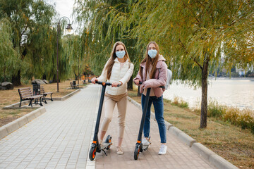 Two young beautiful girls in masks ride electric scooters in the Park on a warm autumn day. Walk in the Park.