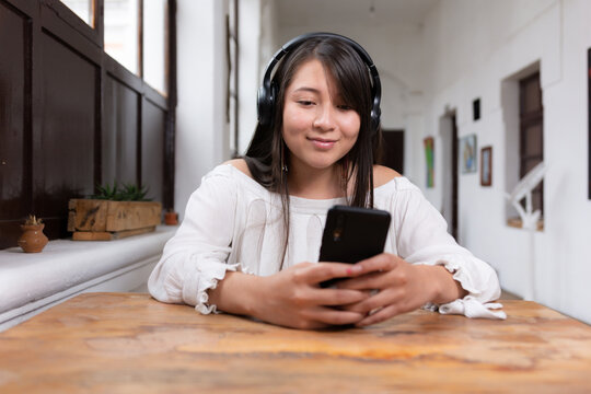 Beautiful happy latina woman with long black hair and wireless headphones listening to music leaning on a wooden table using her cell phone with her hands - Powered by Adobe